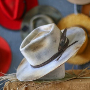 Peut inclure: Un chapeau à larges bords de couleur blanc cassé vieilli avec des nuances gris foncé et marron. Le chapeau présente une bande en cuir marron foncé enroulée autour de la calotte, agrémentée d'une plume sombre glissée sur le côté.