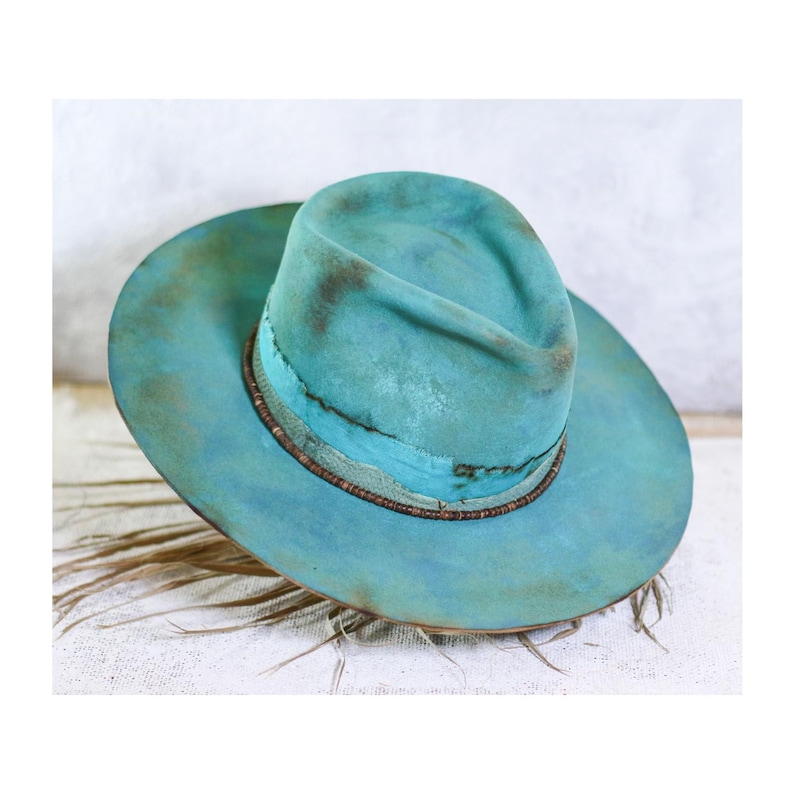 May include: A wide-brimmed hat in a distressed turquoise and teal finish with a pinched crown. The hat features a textured surface with darker brown shading and a thin, dark brown beaded band encircling the base of the crown above the brim.