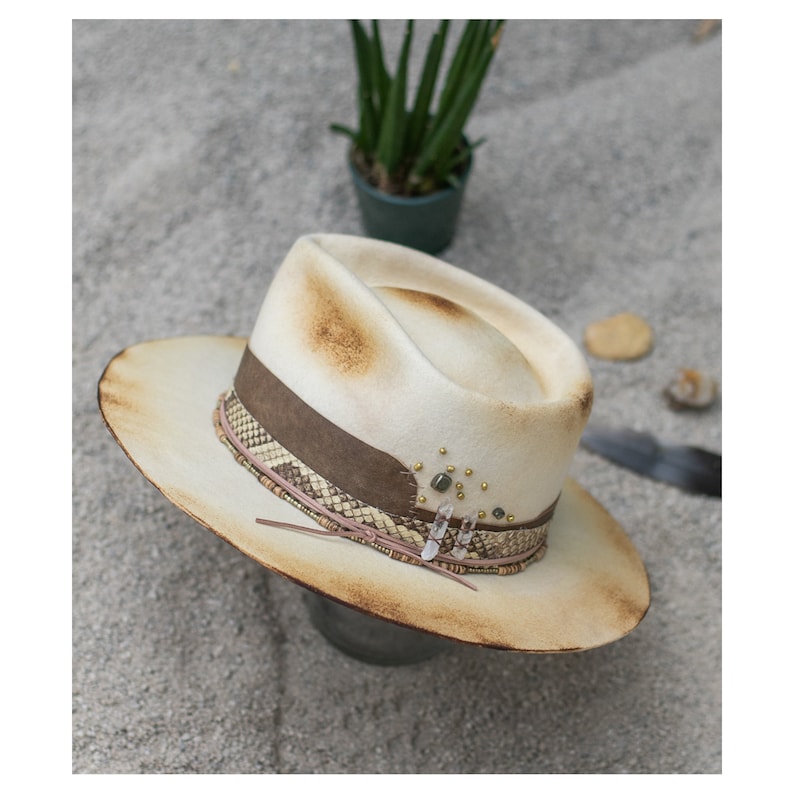 May include: A white felt fedora hat with a brown leather band and a beaded trim. The hat has a distressed look and is decorated with crystals and beads.