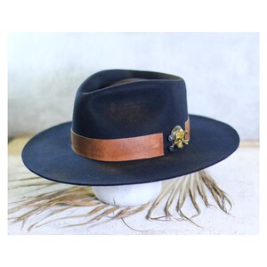 Może przedstawiać: Ciemnogranatowy kapelusz typu fedora z szerokim, gładkim brązowym skórzanym paskiem wokół główki. Metalowy, złoty emblemat w kształcie czaszki jest przymocowany do boku skórzanego paska, stanowiąc charakterystyczny detal tego filcowego kapelusza.