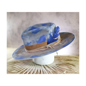 May include: A wide-brimmed hat featuring a grey base with blue painted splotches. The hat is accented with a brown leather band, a thin tan cord, and a single bright blue feather tucked into the side of the crown, creating a distinct visual contrast.