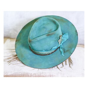 May include: A wide-brimmed hat in a distressed turquoise blue finish with a pinched crown. The hat features a decorative band made of a thin brown cord and a frayed turquoise fabric ribbon tied in a knot on the side of the crown.