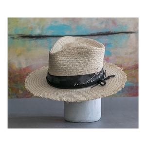 May include: A straw hat with a black ribbon and frayed edges. The hat is sitting on a white pedestal.
