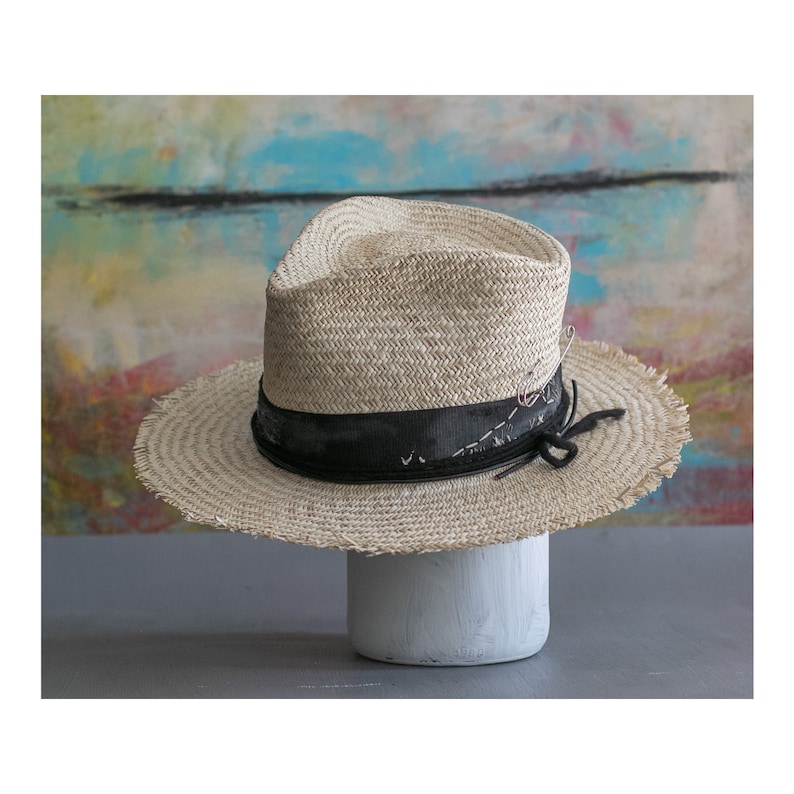 May include: A straw hat with a black ribbon and frayed edges. The hat is sitting on a white pedestal.