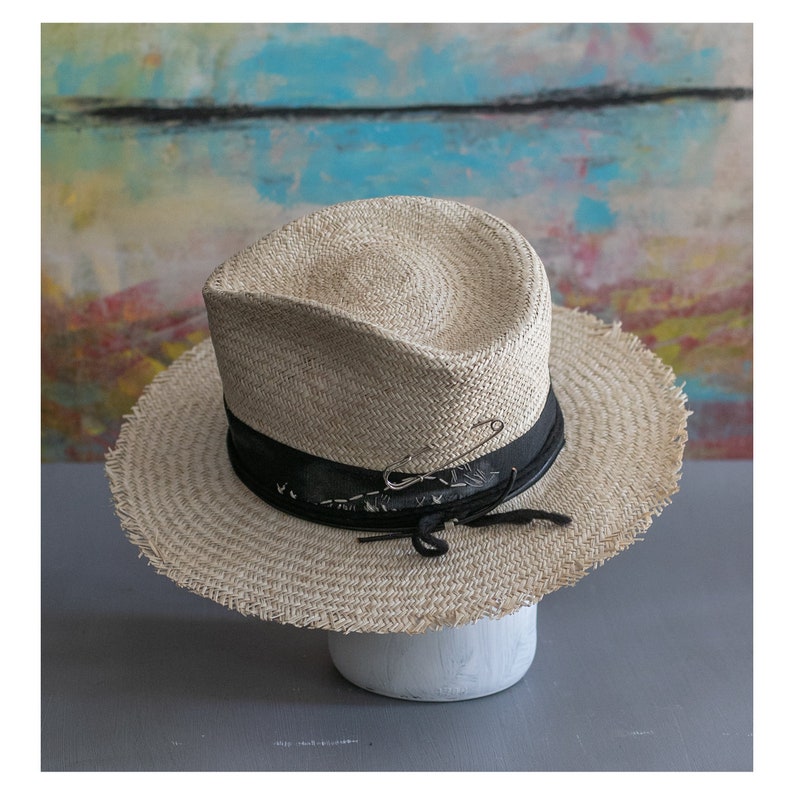 May include: A straw hat with a wide brim and a black band. The hat has a frayed edge and a safety pin attached to the band.