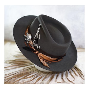 May include: A dark gray fedora hat featuring a brown patterned fabric band, a silver-toned metal skull charm, and a silver-toned safety pin attached to a short metal chain. The hat has a wide brim and a pinched crown design.