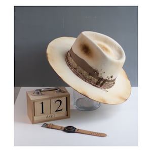 May include: A white felt fedora hat with a brown leather band and gold accents. The hat has a distressed look and is sitting on a glass surface. A wooden calendar with the date "12 Avril" is in the foreground.