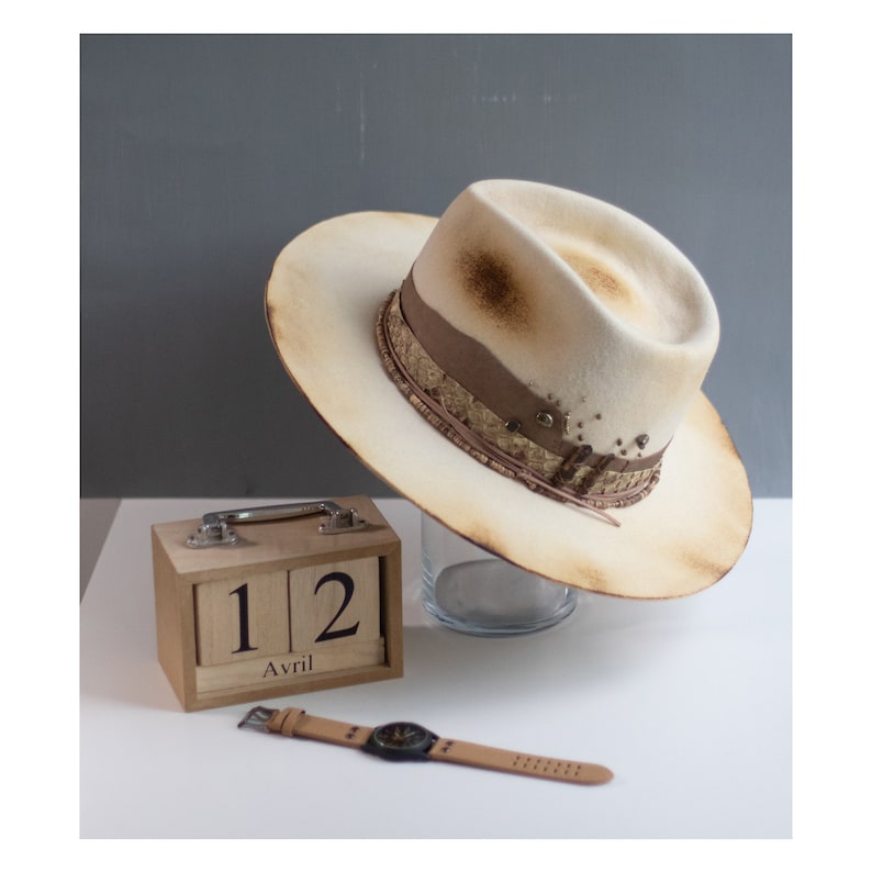May include: A white felt fedora hat with a brown leather band and gold accents. The hat has a distressed look and is sitting on a glass surface. A wooden calendar with the date "12 Avril" is in the foreground.