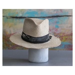 May include: A light brown straw fedora hat with a black ribbon and a frayed edge. The hat is sitting on a white pedestal.