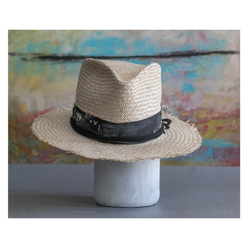 May include: A light brown straw fedora hat with a black ribbon and a frayed edge. The hat is sitting on a white pedestal.