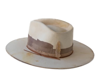 May include: A wide-brimmed off-white hat featuring a distressed finish, a wide taupe band around the crown, a textured woven trim, and a translucent, pointed crystal-like accent attached to the side of the hat band with thin cord.
