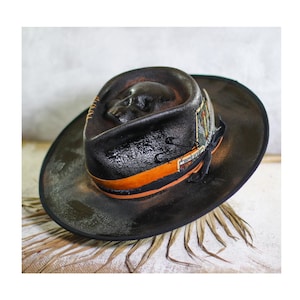 Puede incluir: Un sombrero fedora de color marrón oscuro con un detalle de calavera moldeada en la copa, una cinta naranja y una carta de juego decorativa en el lateral. El ala tiene un acabado desgastado con borde negro y lleva un pequeño imperdible.