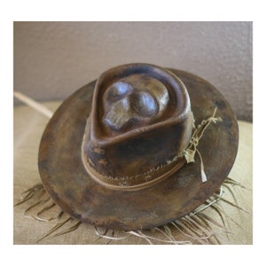 Könnte beinhalten: Ein brauner Fedora-Hut mit einem Totenkopf-Design auf dem Oberteil. Der Hut hat eine breite Krempe und ein dekoratives Band am unteren Rand. Der Hut besteht aus einem strukturierten Material und hat ein verwittertes Aussehen. Rustikaler Stil.