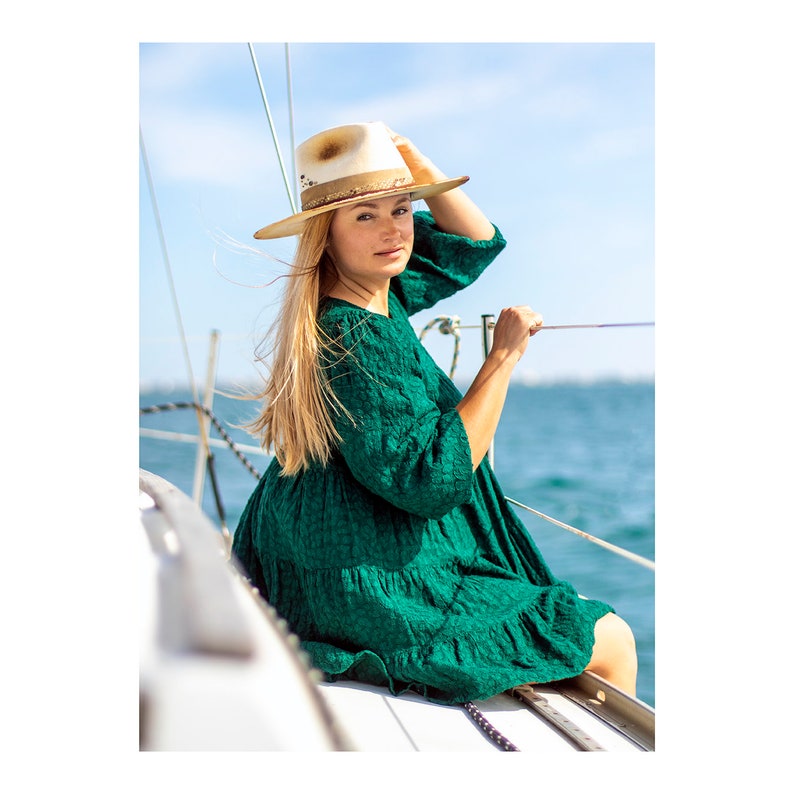 May include: A woman wearing a green dress and a straw hat sits on a boat, looking at the camera. The boat is on the water, and the sky is blue.