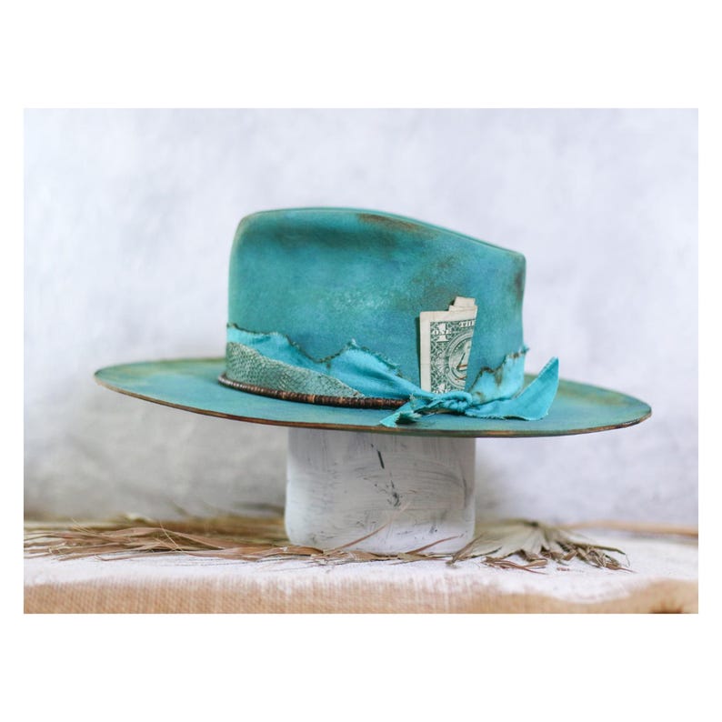 May include: A turquoise fedora hat with a distressed finish, featuring a wide brim and a crown wrapped in a matching fabric band. A small, folded one-dollar bill is tucked into the side of the band, which is accented with a textured, snakeskin-patterned trim.