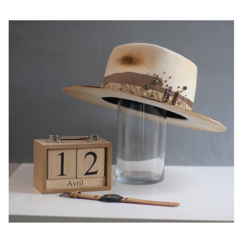 May include: A white felt cowboy hat with a brown leather band and decorative accents. The hat is displayed on a glass stand. A wooden calendar block with the date "12 Avril" is in the foreground.
