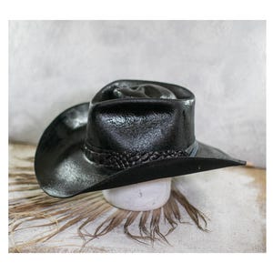 Può includere: Un cappello da cowboy nero con una finitura testurizzata e usurata, caratterizzato da una tesa larga e curva. Il cappello presenta una fascia nera intrecciata che circonda la base della corona, con una piega centrale profonda e lati pizzicati.