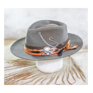 Peut inclure: Un chapeau fedora en feutre gris orné d'une bande en tissu à motifs marron et orange, d'un emblème en métal argenté représentant un crâne et des os croisés, d'une chaîne argentée, d'une grande épingle à nourrice et de plumes sombres.