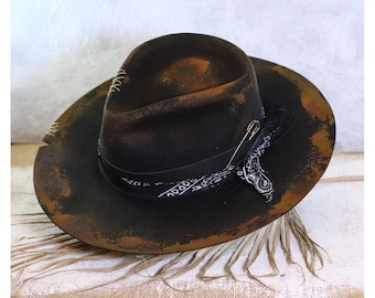 Könnte beinhalten: Ein schwarzer Filzhut im Fedora-Stil mit einem abgenutzten, verwitterten Look. Der Hut hat ein schwarzes Band mit einem weißen Paisley-Muster und eine Sicherheitsnadel.