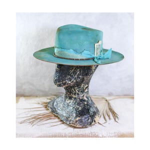 May include: A wide-brimmed teal hat with a distressed finish, featuring a matching teal ribbon band, a thin brown cord, and a small rectangular white tag tucked into the side. The hat is displayed on a dark, textured, abstract head form.