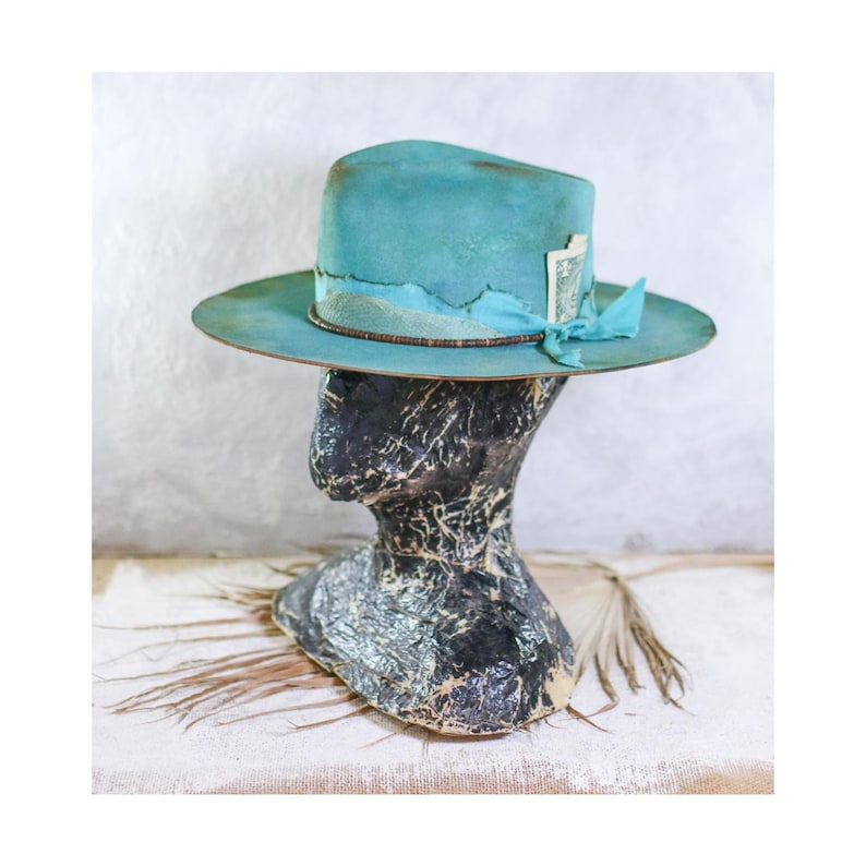 May include: A wide-brimmed teal hat with a distressed finish, featuring a matching teal ribbon band, a thin brown cord, and a small rectangular white tag tucked into the side. The hat is displayed on a dark, textured, abstract head form.