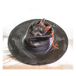 May include: A black fedora hat with a skull design on the crown. The skull has red horns and orange paint splatters on the brim.