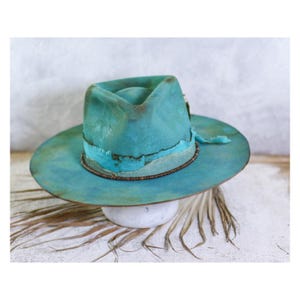 May include: A wide-brimmed hat in a distressed teal and turquoise finish with a pinched crown. The hat features a textured band around the base, accented with a thin brown cord and a frayed fabric trim that adds a layered, worn appearance.