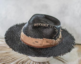 May include: A black straw hat with a wide, frayed brim and a pinched crown. The hat features a tan fabric band wrapped around the base, accented with visible light-colored stitching and small distressed holes in the fabric material.
