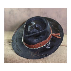 May include: A black leather cowboy hat with a brown leather band and a silver anchor charm. The hat has a distressed look and is decorated with stitching.