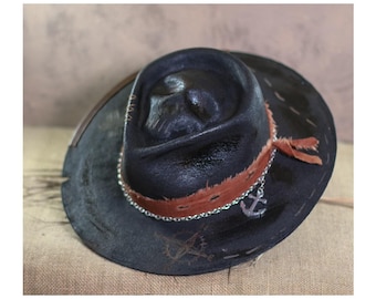 May include: A black leather cowboy hat with a brown leather band and a silver anchor charm. The hat has a distressed look and is decorated with stitching.