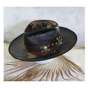 Op de afbeelding: Een zwarte fedora hoed met een verweerde afwerking, voorzien van een donkerbruine leren band versierd met een metalen schedel-embleem. Een zwart lint met gele hartsymbolen en het cijfer zeven is rond de kroon gewikkeld en vastgemaakt met dunne koorden.