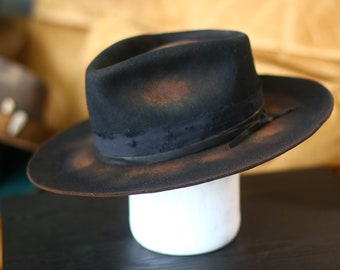 May include: A black felt fedora hat featuring a distressed finish with visible brown patches on the crown and wide brim. The hat includes a dark, frayed fabric ribbon band wrapped around the base of the crown, adding texture to the overall design.