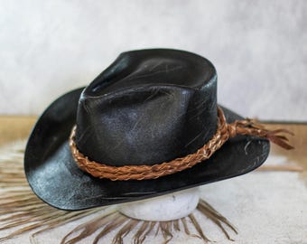 May include: A black cowboy hat with a distressed finish and a textured surface. The hat features a brown braided leather hatband that wraps around the crown and is tied in a knot on the side, creating a distinct contrast against the dark material.