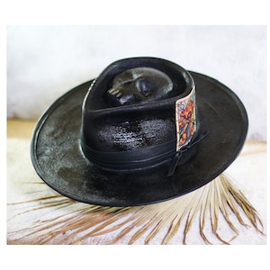 May include: A black fedora hat featuring a moulded skull detail on the crown. The hat includes a black leather band wrapped around the base and a rectangular graphic patch on the side, secured with visible stitching. The brim is wide and flat.
