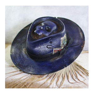 Peut inclure: Un chapeau fedora bleu foncé avec une forme de crâne moulée sur la calotte, un ruban noir et une carte de tarot fixée par une épingle à nourrice noire. Les bords du chapeau présentent des détails usés avec des coutures et des ombres.