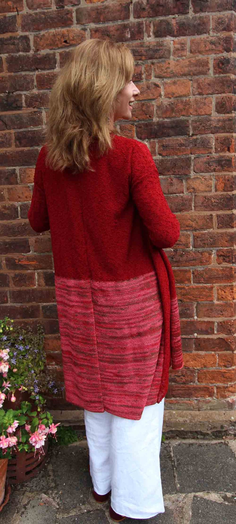 May include: A red and pink knitted cardigan with a long, flowing design. The cardigan is worn over white pants.