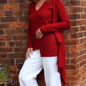 May include: A woman wearing a red knitted cardigan over a red v-neck sweater and white wide-leg pants. She is standing in front of a brick wall and wearing red shoes.