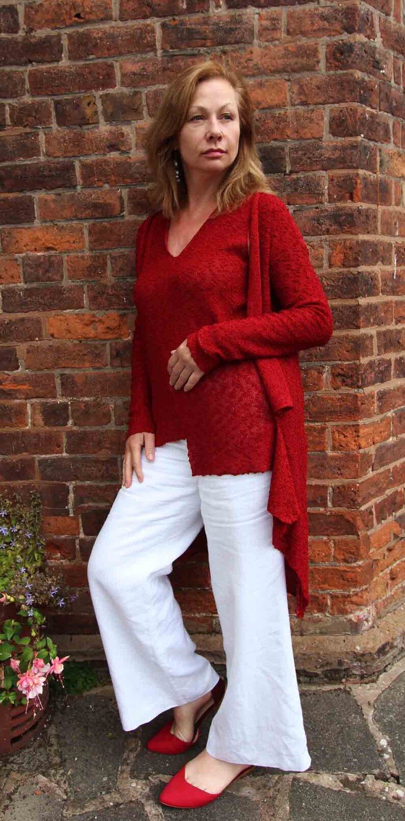 May include: A woman wearing a red knitted cardigan over a red v-neck sweater and white wide-leg pants. She is standing in front of a brick wall and wearing red shoes.