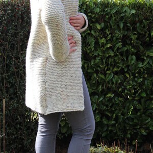 May include: A woman wearing a white knitted cardigan with a speckled pattern and gray jeans. She is standing in front of a green hedge and wearing brown ankle boots.