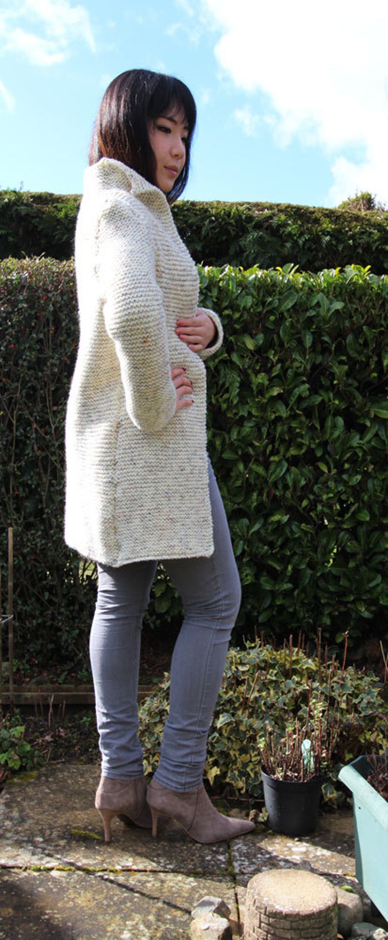 May include: A woman wearing a white knitted cardigan with a speckled pattern and gray jeans. She is standing in front of a green hedge and wearing brown ankle boots.