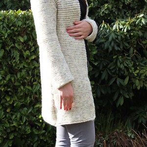 May include: A woman wearing a white knitted cardigan with a long, open front and gray jeans. She is standing in front of a green hedge.