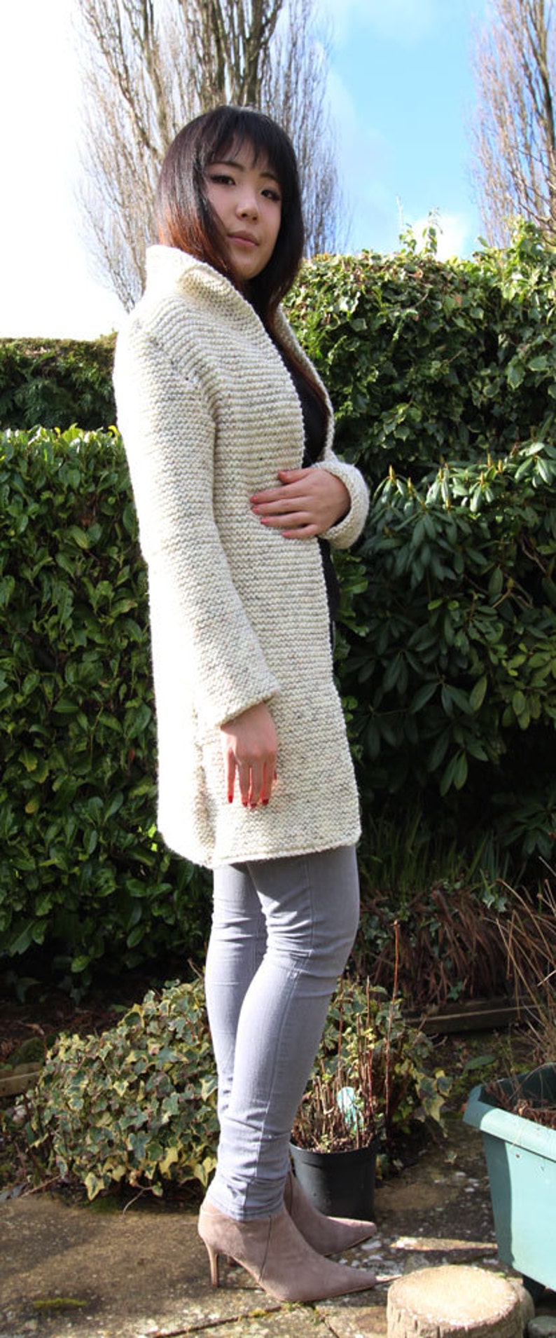 May include: A woman wearing a white knitted cardigan with a long, open front and gray jeans. She is standing in front of a green hedge.