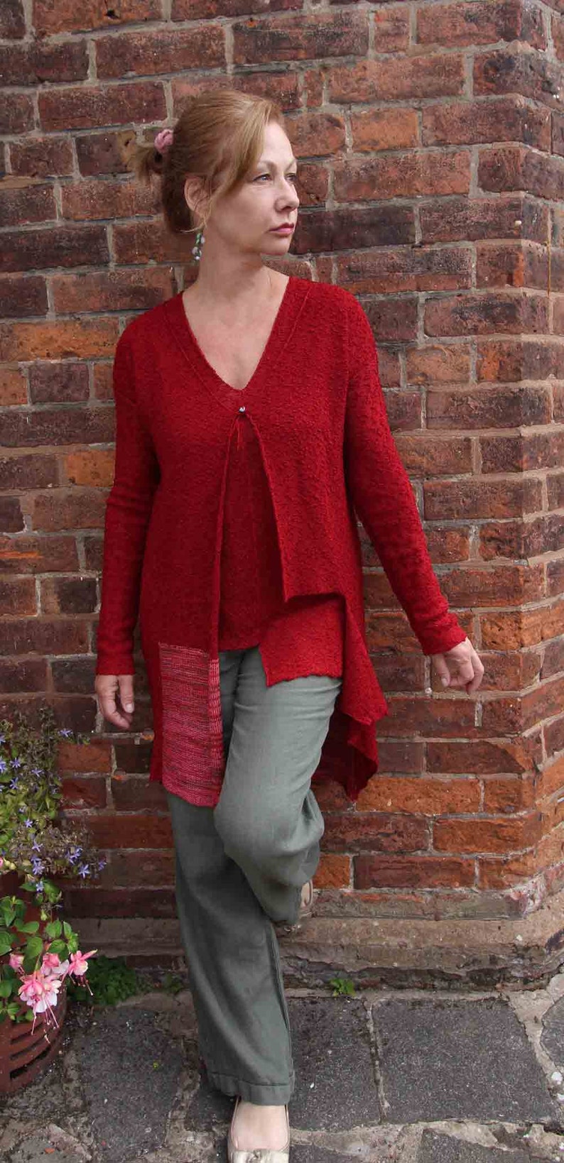 May include: A woman wearing a red, textured cardigan with a long, asymmetrical hemline. The cardigan has a V-neck and a single button closure. She is also wearing green pants.