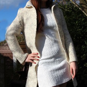 May include: A woman wearing a white knitted cardigan with a white dress underneath. The cardigan has a speckled pattern and a long, open front. The dress has a textured diamond pattern.