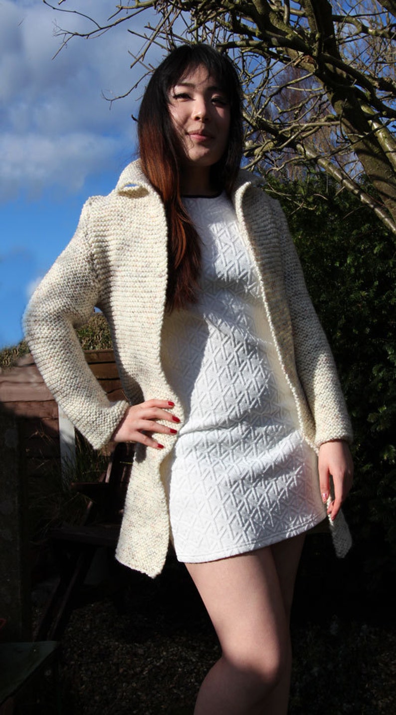 May include: A woman wearing a white knitted cardigan with a white dress underneath. The cardigan has a speckled pattern and a long, open front. The dress has a textured diamond pattern.