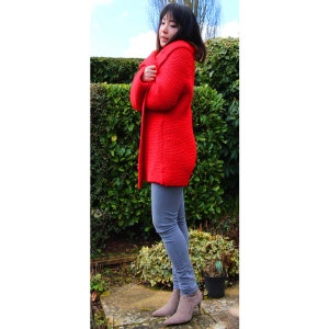 Puede incluir: Una mujer con un c&aacute;rdigan de punto rojo y pantalones vaqueros grises. Est&aacute; de pie frente a un arbusto verde y lleva botines grises.