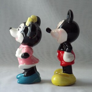 Puede incluir: Dos figuras de cer&aacute;mica de Mickey y Minnie Mouse. Mickey lleva pantalones cortos rojos, guantes blancos y zapatos amarillos. Minnie viste un vestido rosa con lunares blancos, un lazo amarillo y zapatos azules. Ambos tienen orejas negras y caras blancas.