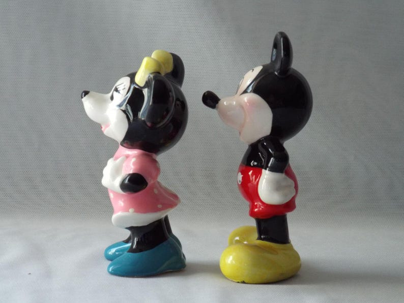 Puede incluir: Dos figuras de cer&aacute;mica de Mickey y Minnie Mouse. Mickey lleva pantalones cortos rojos, guantes blancos y zapatos amarillos. Minnie viste un vestido rosa con lunares blancos, un lazo amarillo y zapatos azules. Ambos tienen orejas negras y caras blancas.