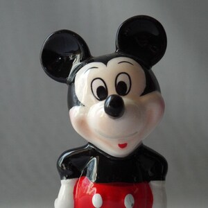 Puede incluir: Una figura de cer&aacute;mica de Mickey Mouse. La figura tiene una cabeza negra con orejas grandes y redondas, una cara blanca y una nariz negra. La figura lleva una parte superior negra con una parte inferior roja y guantes blancos.