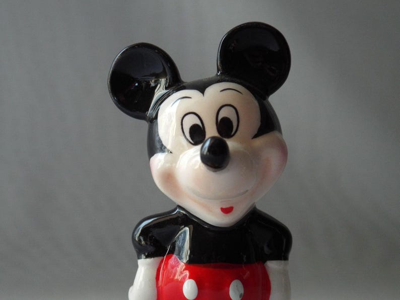 Puede incluir: Una figura de cer&aacute;mica de Mickey Mouse. La figura tiene una cabeza negra con orejas grandes y redondas, una cara blanca y una nariz negra. La figura lleva una parte superior negra con una parte inferior roja y guantes blancos.